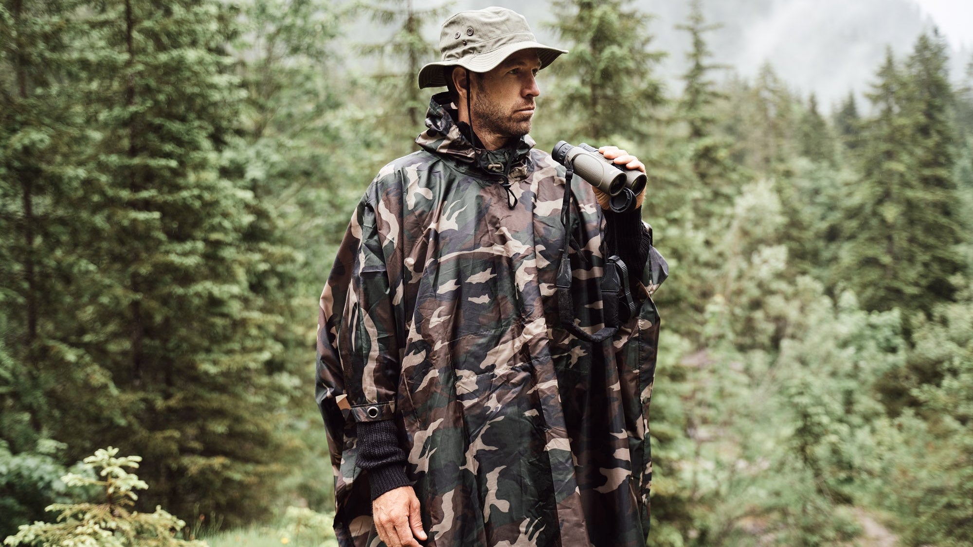A man wearing a camouflage hooded rain poncho and a boonie hat stands in a misty pine forest holding binoculars.