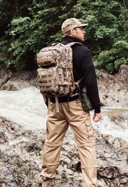 Person wearing a camouflage backpack standing on rocky terrain with a forest background